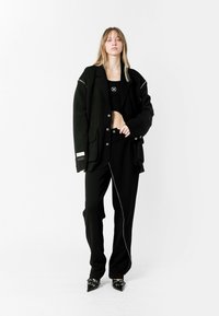 Blazer negro de gran tamaño con acentos plateados, combinado con un top negro corto y pantalones ajustados. Presenta tacones negros de punta. Textura suave.