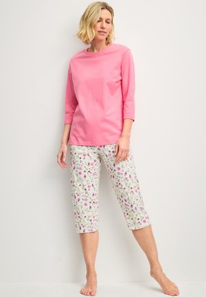 Femme debout pieds nus portant un haut rose à manches trois-quarts et un pantalon blanc court avec un motif floral rose et vert.