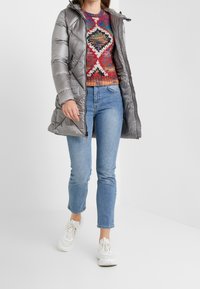 Vrouw in een zilveren pufferjack, gekleurd patroontrui, lichtblauwe jeans en witte sneakers, staand op een witte achtergrond.