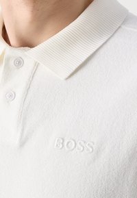 Gros plan d'une personne portant un polo blanc texturé avec col côtelé et deux boutons, logo "BOSS" embossé sur la poitrine.