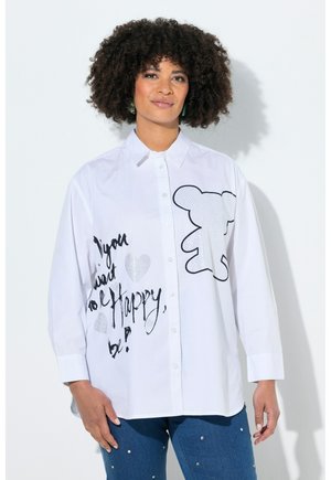 Kobieta o kręconych włosach, ubrana w białą koszulę zapinaną na guziki z czarnym nadrukiem misia i tekstem pisanym odręcznie, w połączeniu z niebieskimi dżinsami ozdobionymi perłowymi aplikacjami.