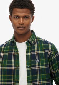 Grün und marinekarierte Flanellhemd mit Knopfleiste, Kragen und aufgenähtem Logo. Getragen über einem weißen T-Shirt, das einen strukturierten Stoff zeigt.