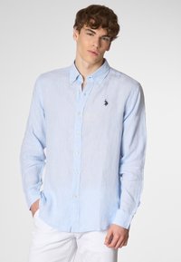 Giovane uomo con capelli scompigliati che indossa una camicia azzurra chiara e pantaloni bianchi, in piedi contro uno sfondo neutro, mentre guarda verso la telecamera.