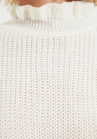 Bílé pletené svetřík s vysokým, volánkovým límcem a otevřenou vazbou. Materiál vypadá měkký a prodyšný.