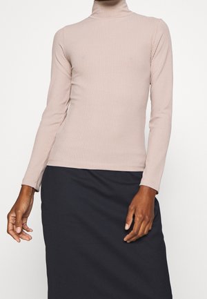 Osoba ubrana w beżowy podkoszulek z długim rękawem i golfem w prążki oraz czarną spódnicę, stojąca na tle jednolitego jasnego tła.