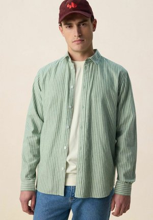 Man draagt een rode pet, een lichtgroen overhemd met verticale strepen over een beige T-shirt en een blauwe spijkerbroek, staand tegen een effen achtergrond.