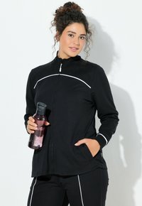 Schwarze Sportjacke mit weißen Akzentnähten, hohem Kragen und Reißverschluss. Das Modell hält eine rosa Wasserflasche und trägt passende Hosen.