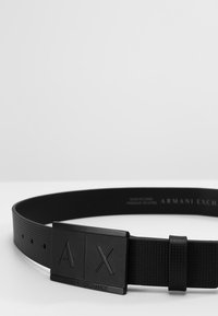 Sorte bælte lavet af tekstureret materiale med en rektangulær spænde, der har et præget "A|X" logo og "ARMANI EXCHANGE" tekst på remmen.
