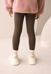 Barna bordázott leggings, rózsaszín plüss bő szabású pulóverrel és krémszínű részletekkel díszített fehér sportcipővel párosítva. A zoknik finoman csíkos mintázatúak.