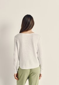 Ženska z dolgimi rjavimi lasmi, oblečena v belo majico z dolgimi rokavi in svetlo zelene hlače, stoji obrnjen proti enobarvni bež steni.