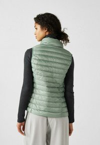 Gilet duveteux vert menthe avec un col haut, design matelassé horizontal et matériau léger, porté sur une chemise à manches longues foncée.