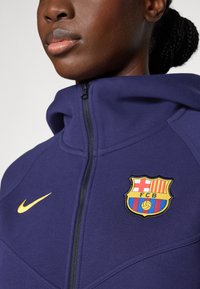 Navyblaue Zip-Hoodie mit hohem Kragen, ausgestattet mit einem gelben Nike-Logo und einem bunten FC Barcelona-Wappen auf der linken Brust. Strukturierten Stoff.