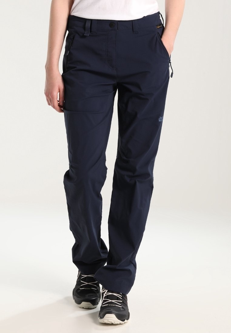 Person wearing navy blue outdoor trousers and black hiking shoes, standing with hands in pockets against plain background.