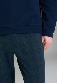 Felpa blu navy con texture liscia; pantaloni a quadri verdi con finitura morbida. Mano visibile appoggiata sui pantaloni.