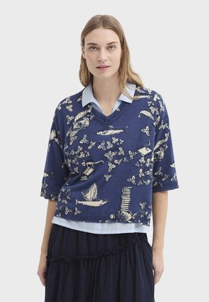 Femme portant un pull bleu à col en V avec des motifs marins et naturels sur une chemise à col bleu clair et une jupe foncée.