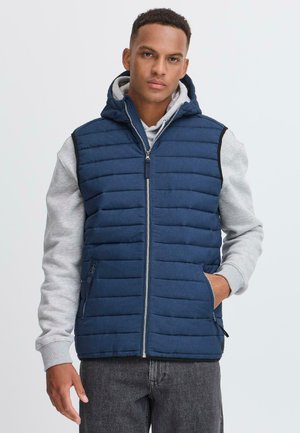Marineblå puffer vest med horisontal quiltet design, glidelås foran og hette. Båret over en grå hettegenser, med sidelommer.