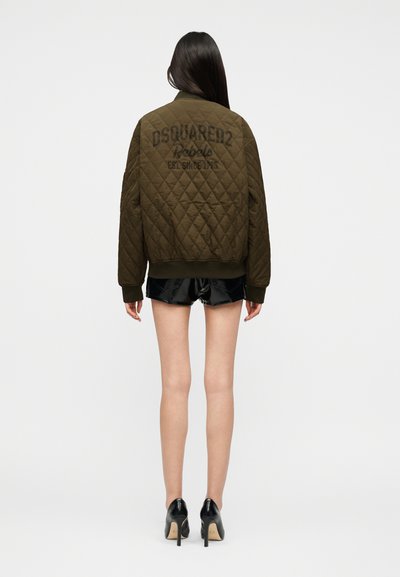 Γυναίκα που φοράει ελιά μπουφάν με το λογότυπο DSQUARED2 Rebels, μαύρα γυαλιστερά σορτς και μαύρα ψηλοτάκουνα, στέκεται με την πλάτη στο λευκό φόντο.