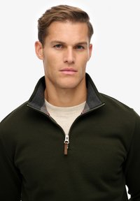 Suéter de color verde oscuro con cuello con cremallera, tejido texturizado y forro interior gris en contraste. Presenta un pequeño acento de cuero cerca de la cremallera.