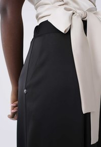 Zwarte broek met een gladde textuur heeft een brede tailleband en een beige satijnen riem die in een strik is geknoopt, met een enkele knoopsluiting.