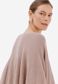 Světle růžový oversized svetr z jemného, texturovaného pletení. Široký výstřih a splývavý design, model nosí texturované zlaté náušnice.