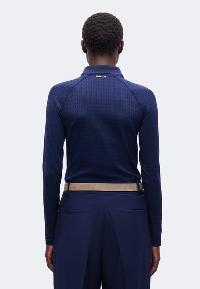 Personne aux cheveux courts et bouclés vue de dos portant un haut texturé bleu marine à manches longues et un pantalon bleu marine avec une ceinture beige.