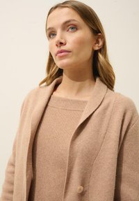 Cárdigan marrón claro con cuello estilo chal, que cuenta con un cierre de un solo botón y una tela suave y texturizada. Lleva puesto un suéter de canalé a juego.