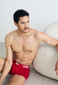 Red Lacoste boxer briefs with white waistband logo, fitted design, and a soft cotton texture. Model seated, with a light background and plush seating.