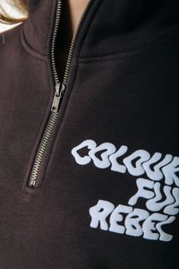 Kratki prikaz crne zip-up dukserice s bijelim stiliziranim tekstom "Colourful Rebel" na prsima.