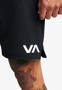 Sorte atletiske shorts med en glat tekstur, der har et hvidt "VA" logo nær kanten og sideslids for bevægelsesfrihed.