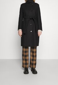 Sort uldfrakke med høj krave, knaplukning og bælte detalje. Kombineret med brune og sorte ternede brede bukser og sorte støvler.