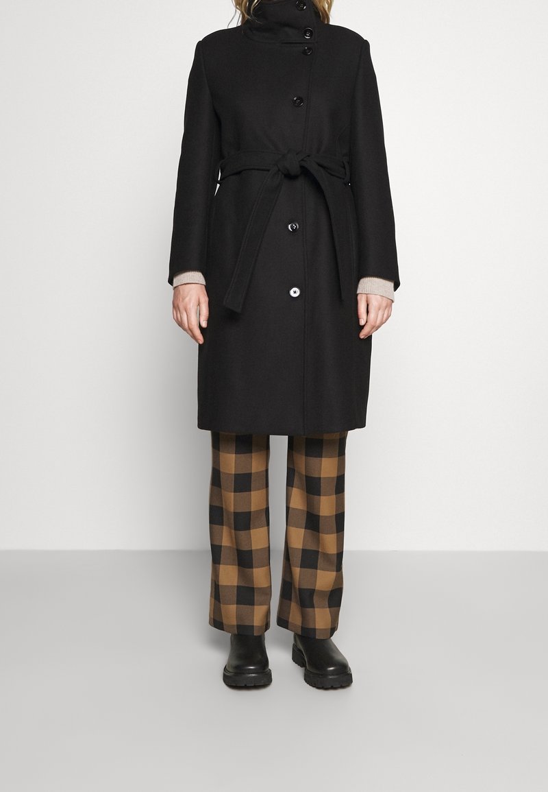 Sort uldfrakke med høj krave, knaplukning og bælte detalje. Kombineret med brune og sorte ternede brede bukser og sorte støvler.