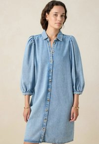 Jeune femme portant une robe en denim bleu clair à boutons avec des manches bouffantes trois-quarts et des bracelets en or aux deux poignets.