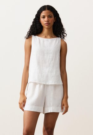 Kvinde med krøllet sort hår iført en ærmeløs hvid hør top og matchende hvide hørshorts mod en ensfarvet lys baggrund.