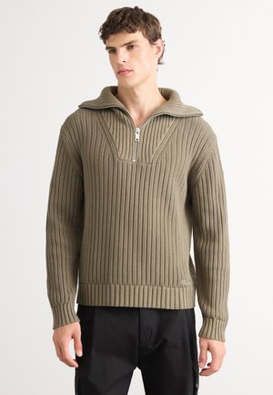 Olive green ribbed sweater with a zip collar, featuring a textured design and slightly fitted silhouette, paired with black pants.