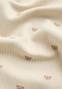 Țesătură crem deschis cu nervuri subtile, având un model repetitiv de fețe mici de urs maro. Moale, texturată și elastică.