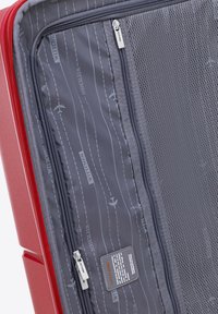 Interior de maleta rígida roja con tela gris que presenta patrones de aviones y carreteras, múltiples compartimentos con cremallera y un separador de malla.