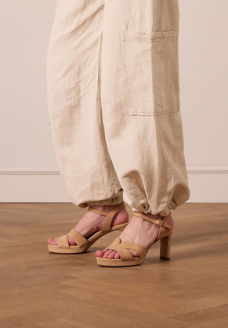 Beige suède sandaaltjes met hak en een strappy ontwerp en open teen. Voorzien van een blokhak en enkelbandje, gecombineerd met lichte linnen broek.