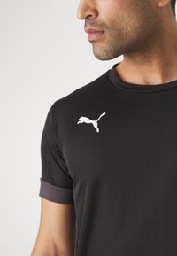 Camisola desportiva preta com decote redondo e mangas curtas, apresentando um detalhe cinza escuro nas bainhas. Exibe um logo branco da Puma no peito.