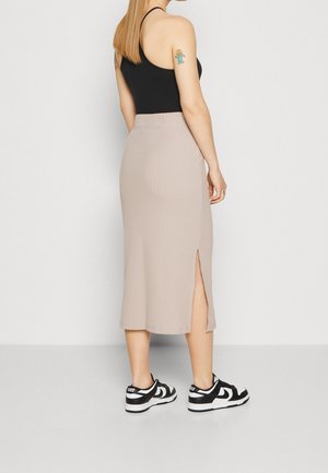 Vrouw draagt een zwarte mouwloze top, beige geribde midi rok met zijsplit en zwart-witte Nike-sneakers, staand zijwaarts.