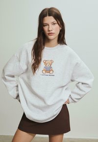 Grå sweatshirt med oversized pasform, prydet med en teddy bear-illustration og teksten "Jeg kan ikke klare mandage". Matches med en brun mini-nederdel.