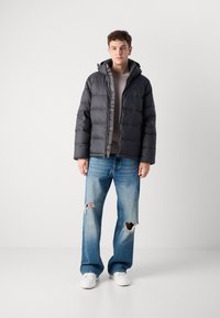 Zwarte pufferjack met een capuchon, met een gewatteerde textuur en een rits aan de voorkant. Draagt over een licht vest en gecombineerd met versleten blauwe jeans.