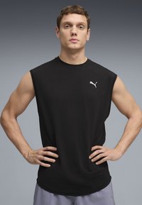 Maglietta sportiva nera senza maniche con scollo rotondo e logo Puma bianco sul lato sinistro del petto; indossata con pantaloncini grigio chiaro.