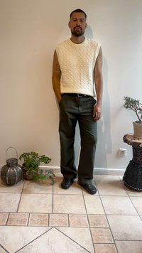 White cable-knit sleeveless sweater, dark green pants, black shoes; standing in a room with plants and decorative items.