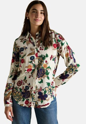 Woman wearing a cream button-up shirt with colorful floral patterns and blue jeans, posing with one hand on hip against a plain background.