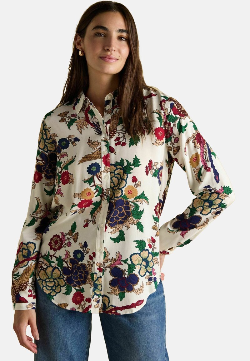 Woman wearing a cream button-up shirt with colorful floral patterns and blue jeans, posing with one hand on hip against a plain background.