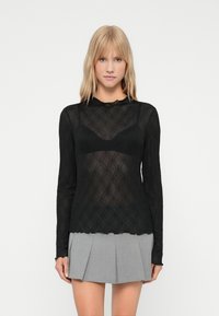 Top negru cu mânecă lungă, având un design transparent și texturat cu un model floral și un decolteu în valuri, asortat cu o fustă plisată gri.