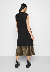 Svart stickad ärmlös tunik med avslappnad passform, lager-på-lager över en veckad olivgrön kjol, ihop med svarta ankelstövlar.