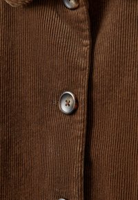Gros plan d’un tissu en velours côtelé marron avec des côtes verticales et de gros boutons ronds cousus en ligne verticale.