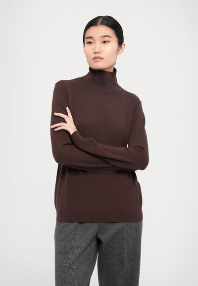 Pull encolure fendue marron en tissu doux avec un design ajusté. Le modèle porte un pantalon à chevrons gris, les bras croisés.