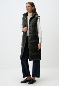 Gilet long noir matelassé avec un col montant, fermeture à pression sur le devant et poches latérales, associé à un pull col roulé crème et un pantalon en denim foncé.